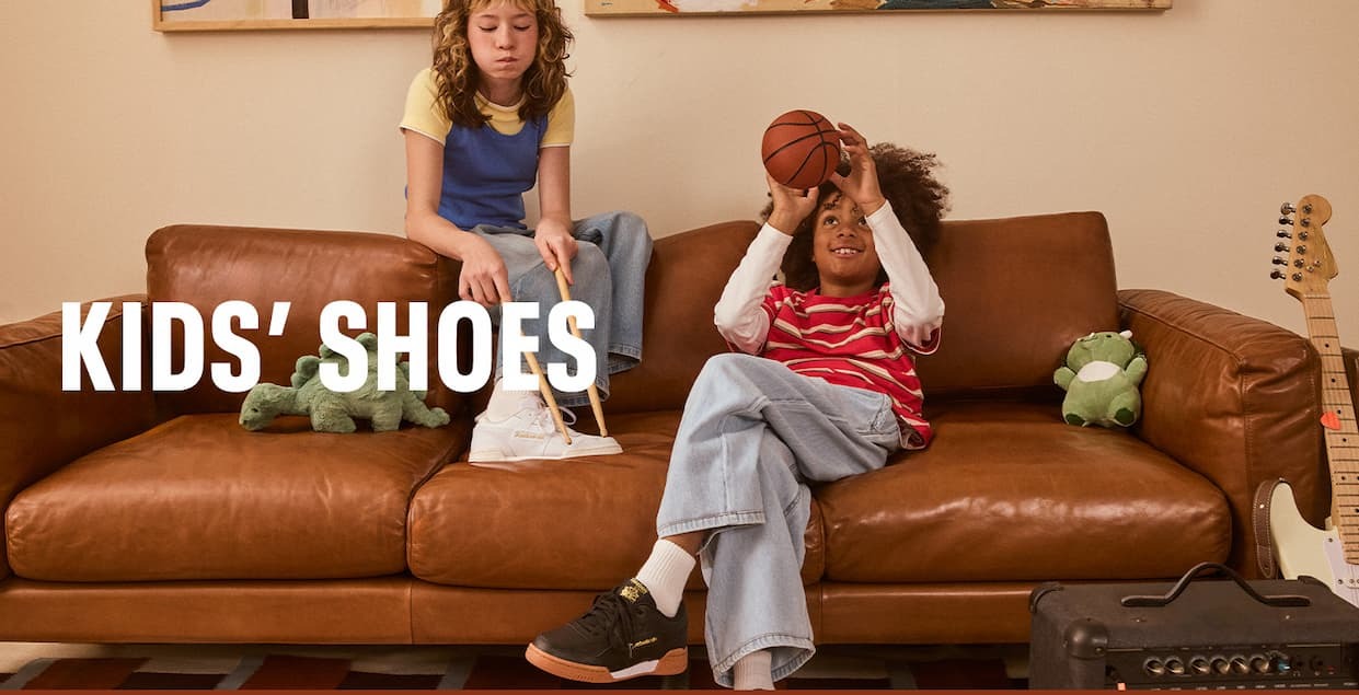 Two kids wearing Reebok kids’ sneakers relax on a couch with a basketball and drumsticks. Text reads: “KIDS’ SHOES.”