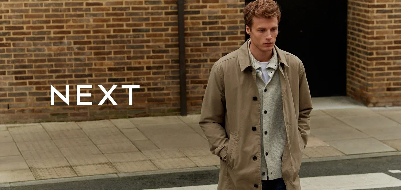 Beige trenchcoat over een grijs sweater en een wit shirt, donkere jeans, wandelend op een zebrapad. Bakstenen muur op de achtergrond, casual stedelijke omgeving.