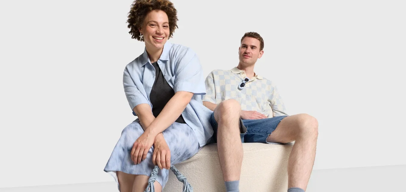 Twee personen zitten op een textuur, ronde, beige poef. De ene draagt een lichtblauw shirt, een spijkerrok en blauwe gestikte ballerina's; de ander draagt een shirt met patroon en spijkershorts.