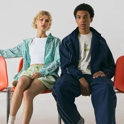 Twee mensen zitten op oranje plastic stoelen. De ene draagt een groen ontworpen jasje en shorts; de ander een marineblauwe trainingspak. Sneakers en sokken zijn zichtbaar.