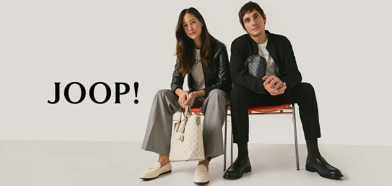 Twee volwassenen zitten op rode stoelen tegen een effen achtergrond; de vrouw houdt een beige handtas vast, de man een zwart patroonpochet, beiden dragen casual kleding.