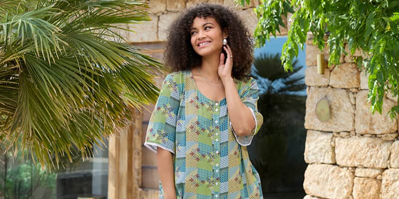 Ein Model in einer Urlaubsumgebung trägt eine ecru-pistazie-bedruckte Tunika mit Halbarm-Flügelärmeln, Alloverdruck und aufgesetzter Knopfleiste am Rundhalsausschnitt