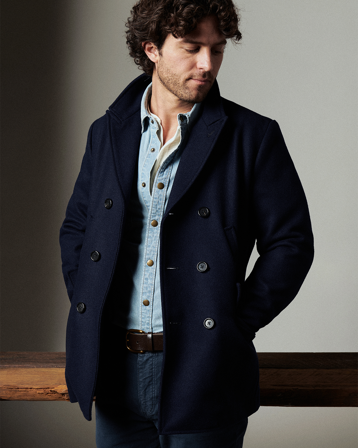 Man with dark curly hair wears a navy blue peacoat with black buttons over a denim shirt.