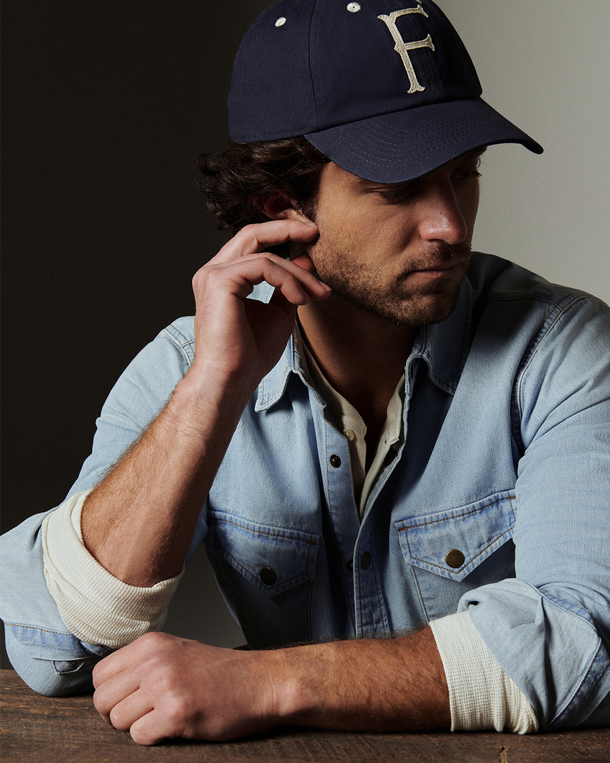 Man with curly dark hair wears a blue baseball cap with a denim shirt while sitting at a table.