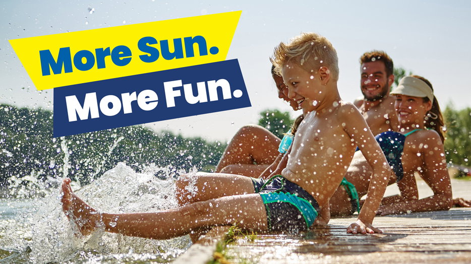 More Sun. More Fun. A group of people sitting along the edge of a beach area with water splashing around their feet on a sunny day.