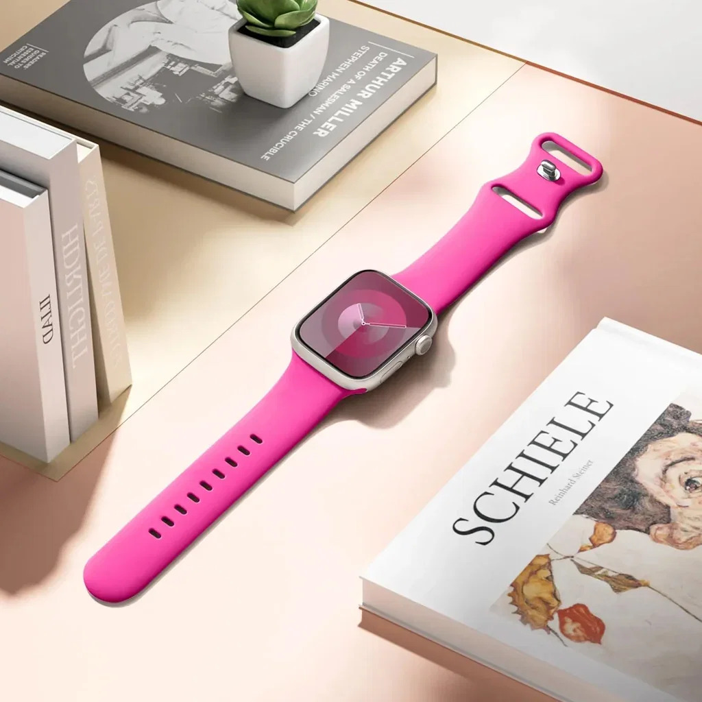 Bright pink smartwatch with matching band displayed on a desk with books and a succulent plant.