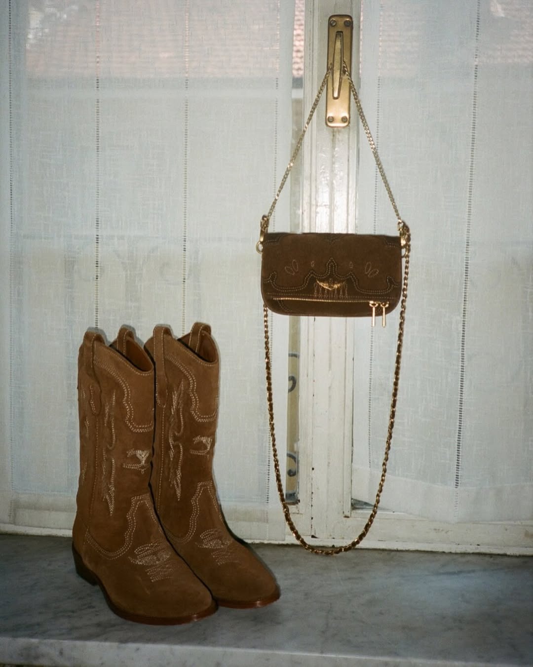 Pair of brown suede cowboy boots on a marble windowsill and a matching brown suede crossbody bag with gold chain hanging from a window latch, sheer curtains behind