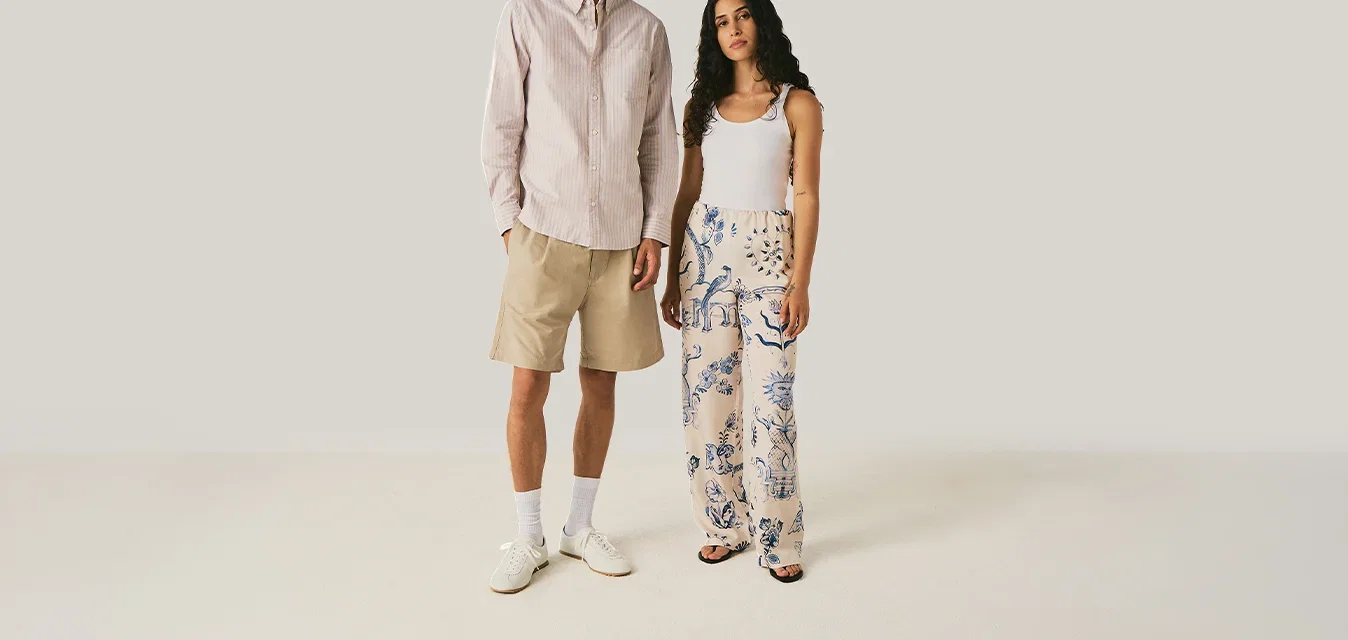 Homme portant un short beige, une chemise blanche, des chaussettes blanches et des chaussures se tenant à côté d'une femme en débardeur blanc et pantalon large à motifs sur fond uni.