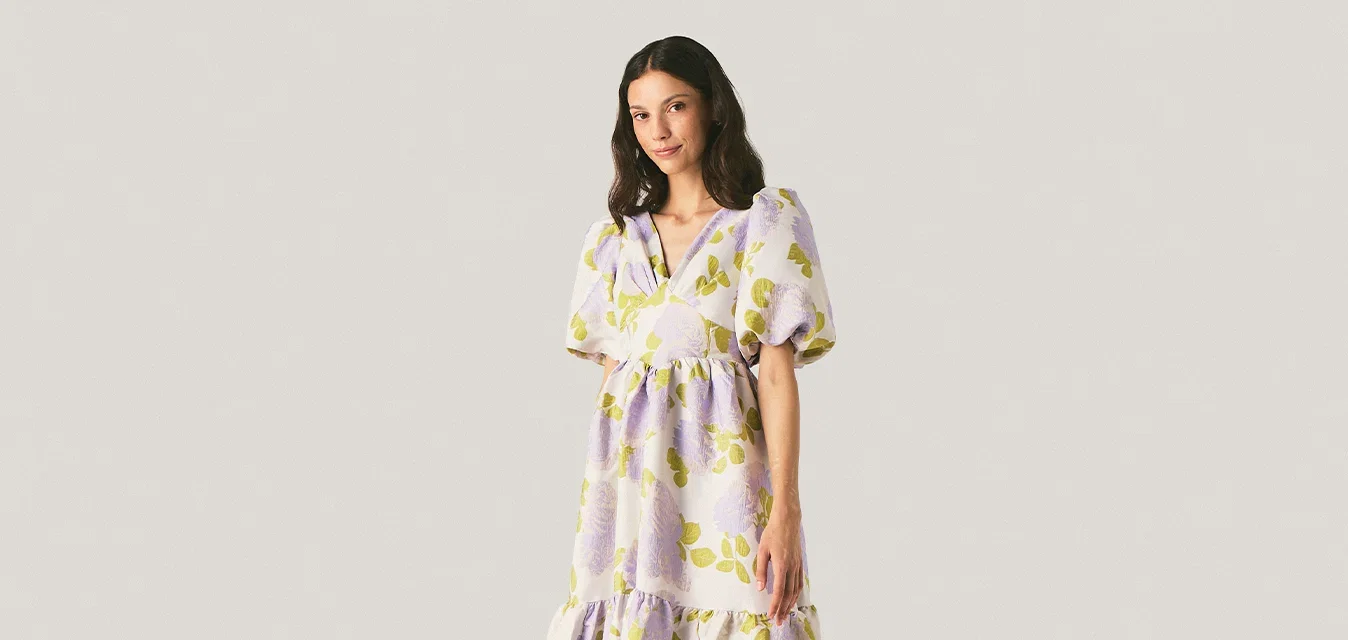 Femme debout dans une robe ample à fleurs avec des manches bouffantes sur un fond clair uni, face à la caméra, les mains détendues le long du corps.
