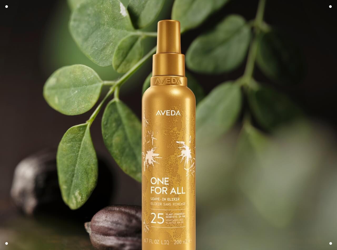 Close-up of a bottle of Aveda One For All Leave-In Elixir surrounded by moringa seeds and leaves.
