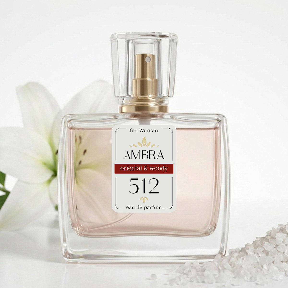 Glass perfume bottle labelled 'AMBRA 512 oriental & woody' beside a white lily and sea salt crystals, eau de parfum