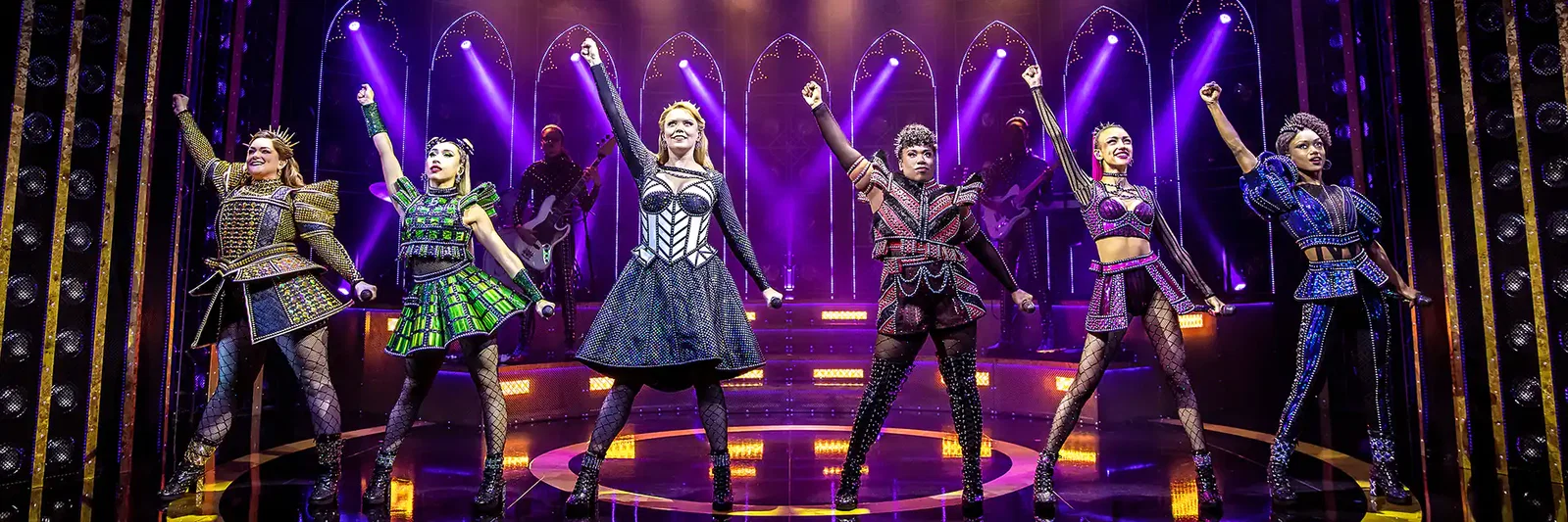 A group of five performers in vibrant costumes striking powerful poses on stage with dramatic lighting.