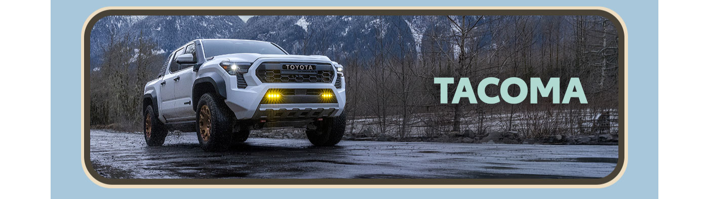 Tacoma. A white Tacoma cruises down a rainy road at dusk. Snowcapped mountains loom in the background. Explore Tacoma.