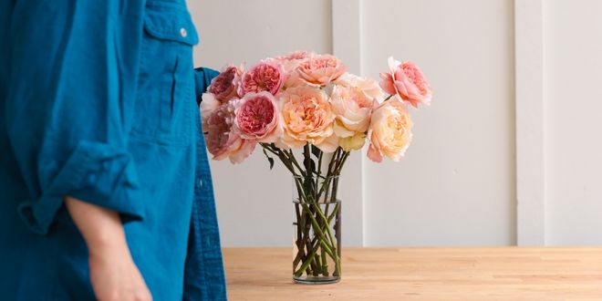 A glass vase of pink and peach garden roses on a wood table as someone in a denim shirt is arranging roses