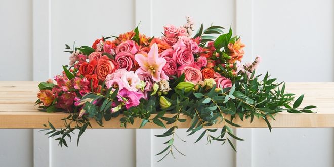 A long, low arrangement of coral and pink roses, a pink lily, and trailing greenery laid across a wood bench.