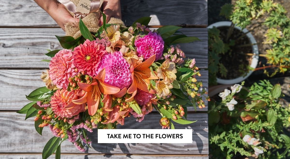 Overhead view of hands holding a burlap-wrapped bouquet of dahlias and orange lilies on a weathered wood deck.