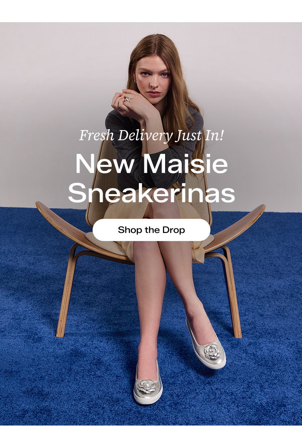 A woman sitting in a wooden chair on a blue carpet. She is wearing a grey long-sleeved shirt and a beige skirt paired with the new Maisie Ballet Metallic Leather Flower Appliqué flats in silver. Text over the image says Fresh Delivery Just In! New Maisie Sneakerinas. Shop the Drop.