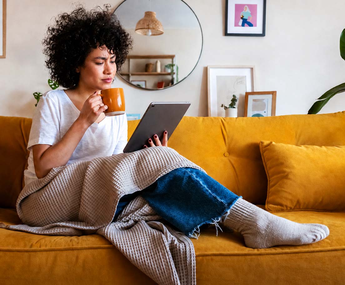 A woman sits on a yellow sofa with a mug and tablet, looking thoughtfully at the screen