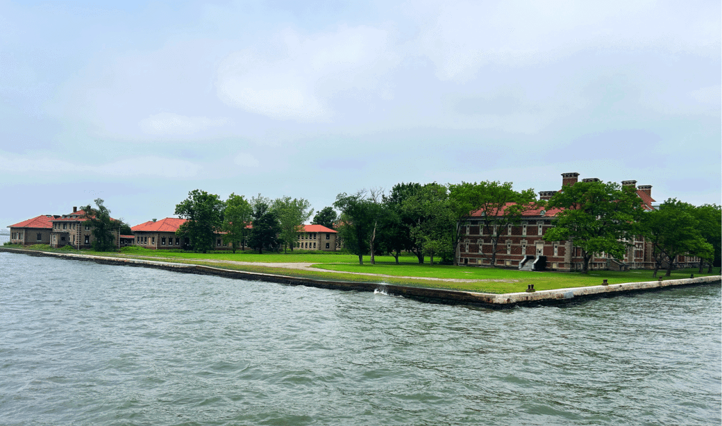 Historic brick buildings with red roofs surrounded by trees sit on a grassy area next to the water under a cloudy sky.