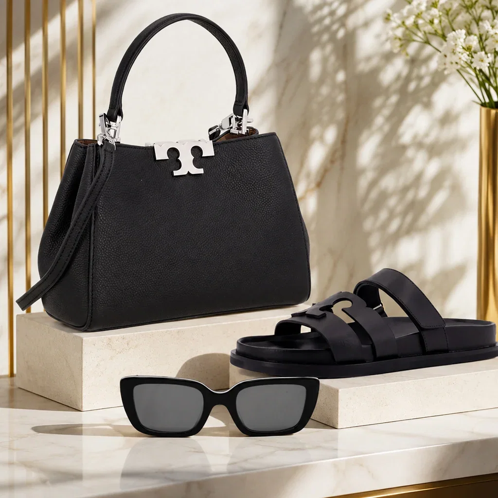 Black leather handbag, sunglasses, and slide sandals displayed on marble platform with gold accents and white flowers.