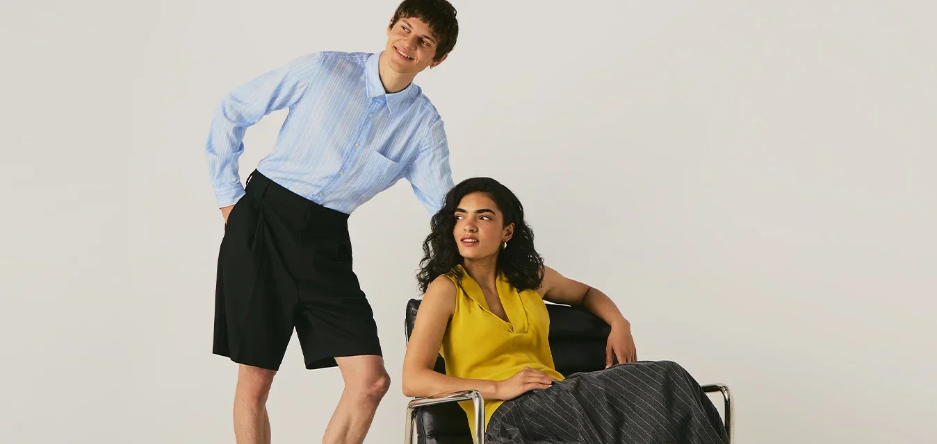 Dos personas con ropa moderna, una sentada en una silla con estructura metálica y la otra de pie a su lado en un espacio neutral.