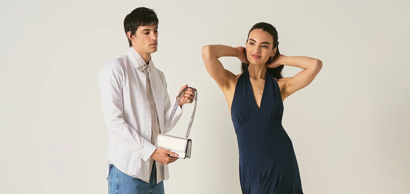 Hombre con camisa blanca y vaqueros sosteniendo un pequeño bolso junto a una mujer con un vestido azul marino que posa con las manos detrás de la cabeza.