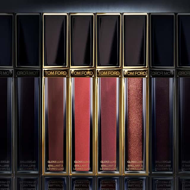 Glossy Tom Ford lip gloss tubes in various shades, including dark red, light pink, and glittery maroon, are arranged in a row under a spotlight. The gold and black packaging adds a luxurious touch.
