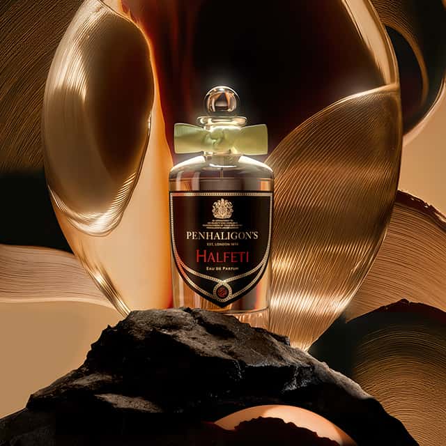 A bottle of Penhaligon's Halfeti Eau de Parfum is placed against a luxurious backdrop featuring gold and bronze swirling patterns. The bottle is elegantly designed with a label reading 'Halfeti,' and a bow adorns the neck of the bottle, enhancing its opulent appearance.