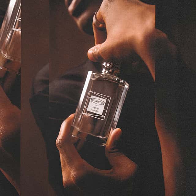 A person holds a glass bottle of perfume labelled 'Be Wanted.' The scene is set in warm lighting with the person's hand softly gripping the bottle. The background is dark, drawing focus to the perfume.