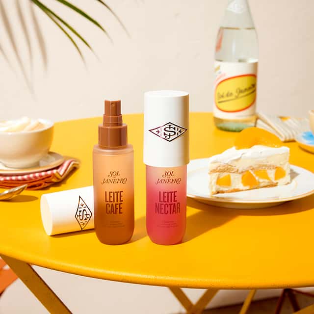 Two Sol de Janeiro hair care products, Leite Café and Leite Nectar, are on a bright yellow table. A plate with a slice of dessert and a bottle in the background. A plant casts a shadow on the wall, creating a summery atmosphere.