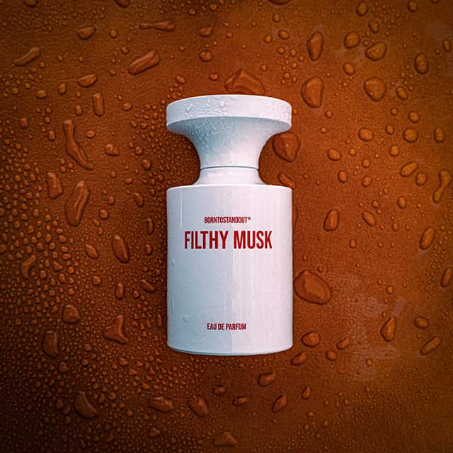 A white perfume bottle labelled 'Filthy Musk Eau de Parfum' stands on a wet, brown surface with water droplets scattered around.