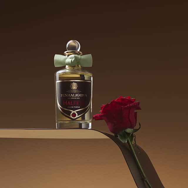 A bottle of Penhaligon's Halfeti perfume with a light green ribbon on its neck is placed on a reflective surface. Beside the bottle lies a single red rose. The background is a warm, brown gradient.