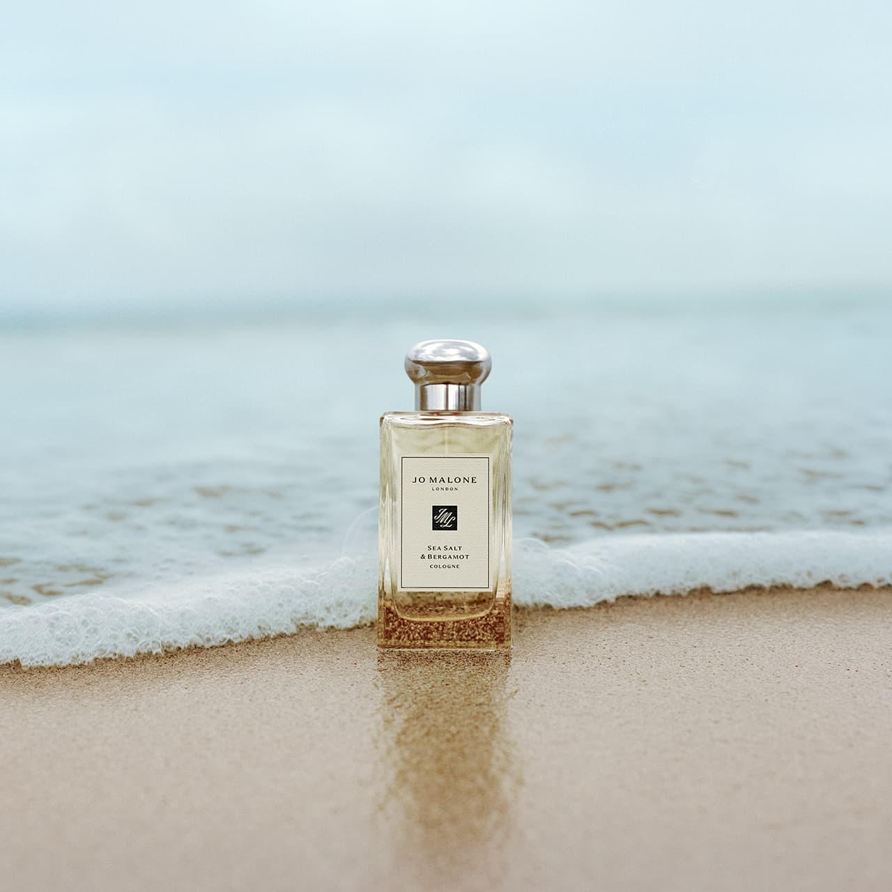 A bottle of Jo Malone perfume stands on wet sand as gentle waves from the sea approach in the background, creating a serene and fresh atmosphere.