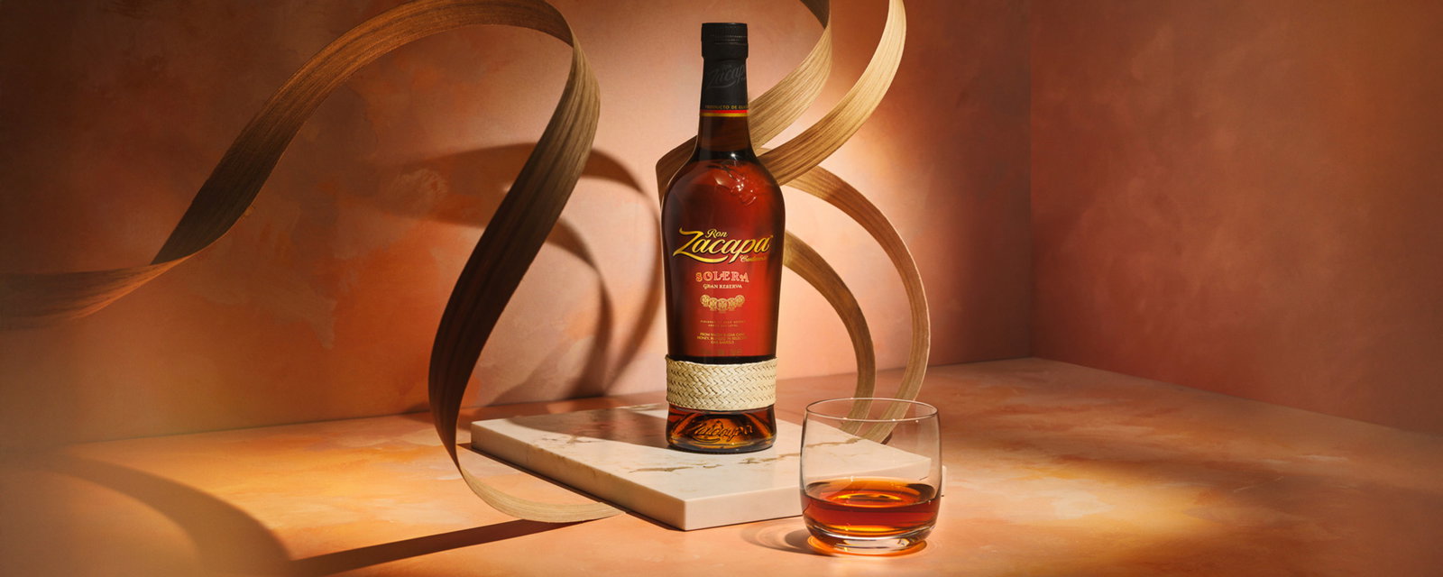 Bottle of Zacapa rum on a marble slab with a glass of amber rum, decorative wooden ribbons and warm, moody lighting