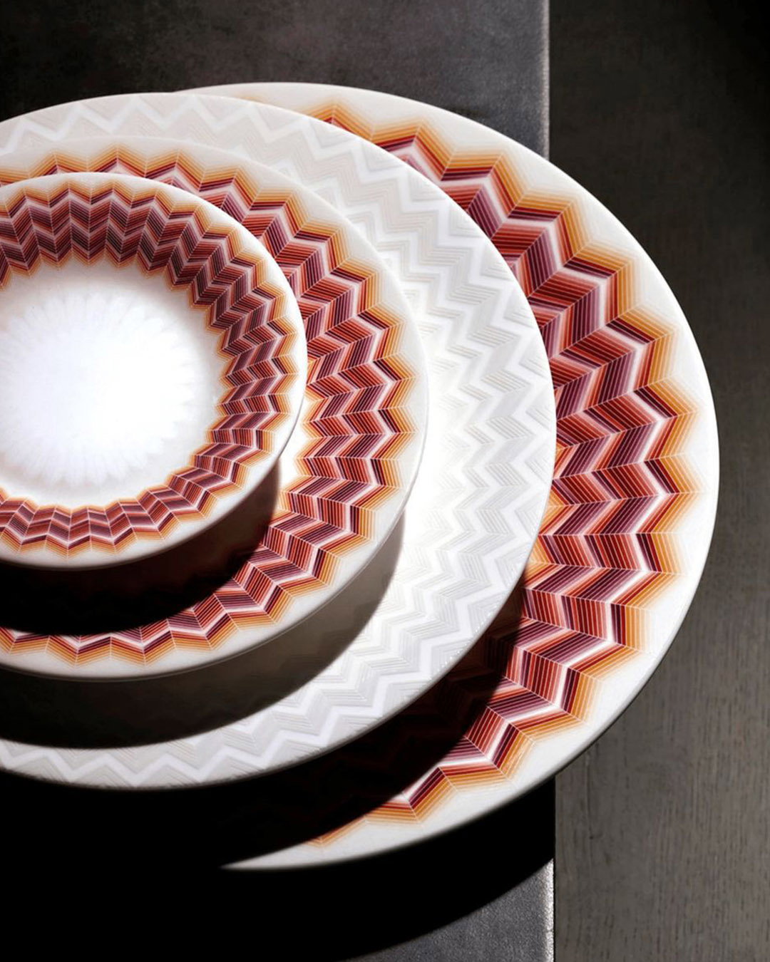 Stack of three white ceramic plates with concentric red‑orange zigzag geometric rims, photographed top‑down on a dark wooden table