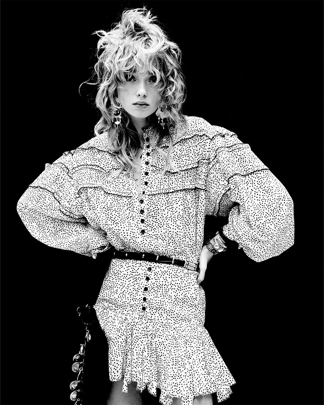 Black-and-white studio portrait of a woman with a shag haircut wearing a polka-dot ruffled blouse and matching skirt, hands on hips, statement earrings and belt