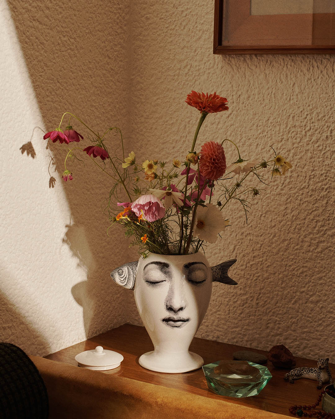 Decorative face vase with fish-shaped handles holding wildflowers on a wooden table in warm sunlight