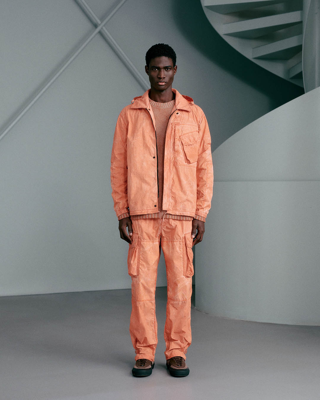 Male model wearing orange utility jacket and cargo trousers, standing in studio with spiral staircase