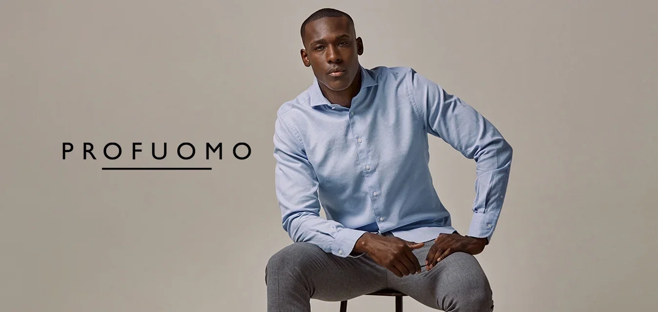 Lichtblauw button-up shirt, grijze broek en grijze sneakers. Model zit op een zwarte kruk met de handen op de knieën tegen een neutrale achtergrond.