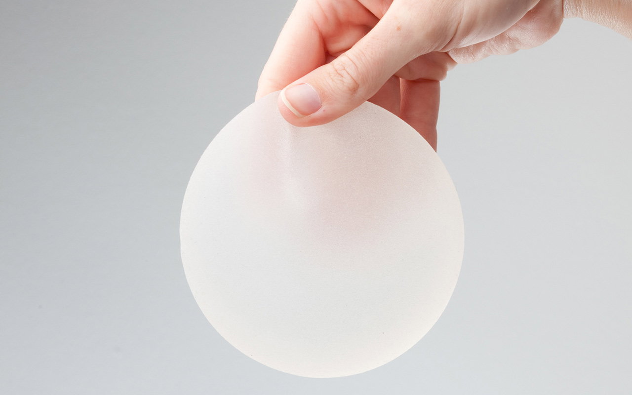 A hand gently holds a round silicone breast implant against a plain gray background