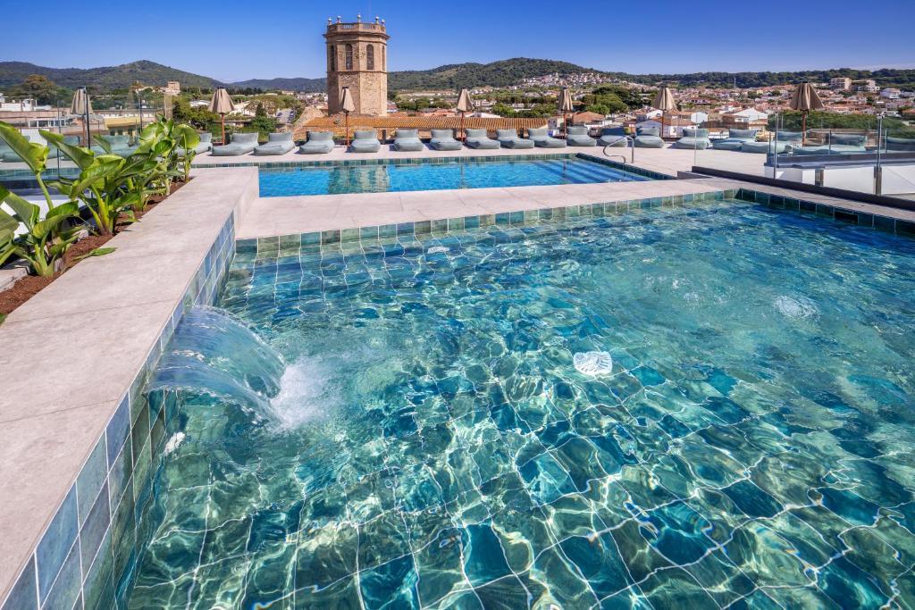Rooftop pools with clear turquoise water, loungers and umbrellas, plus a historic bell tower overlooking a coastal town, hills