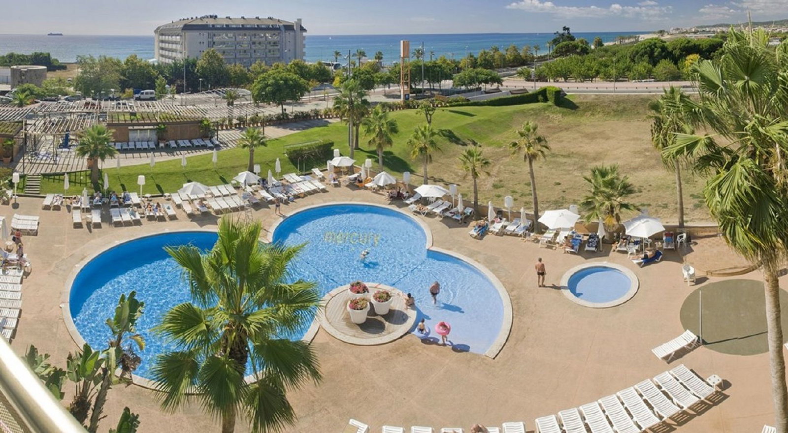 Aerial view of a three-lobed resort swimming pool with sunloungers and palm trees, guests relaxing and the sea visible in the background