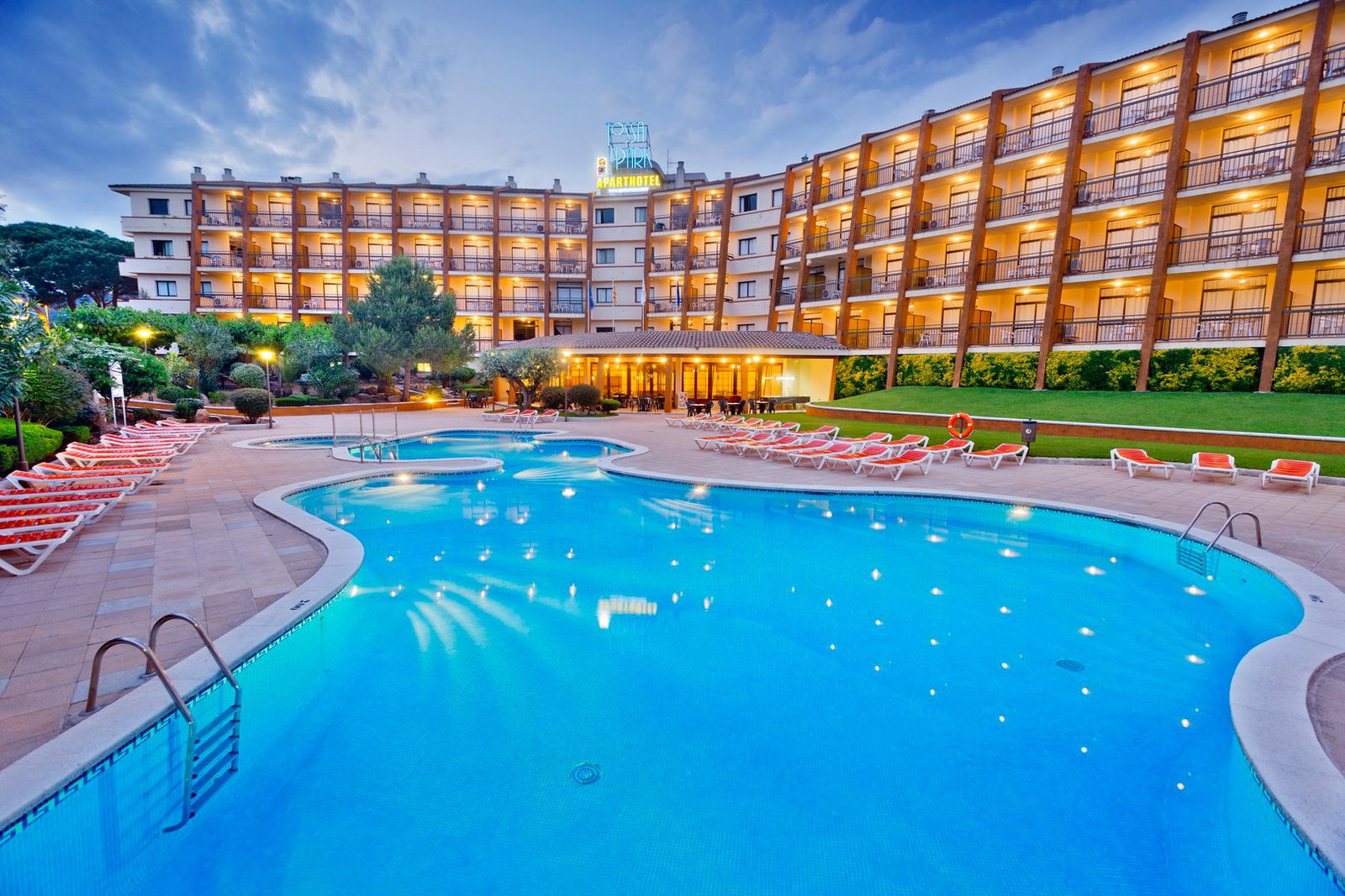 Evening view of a lit aparthotel with a large curved outdoor swimming pool and rows of sun loungers on the terrace