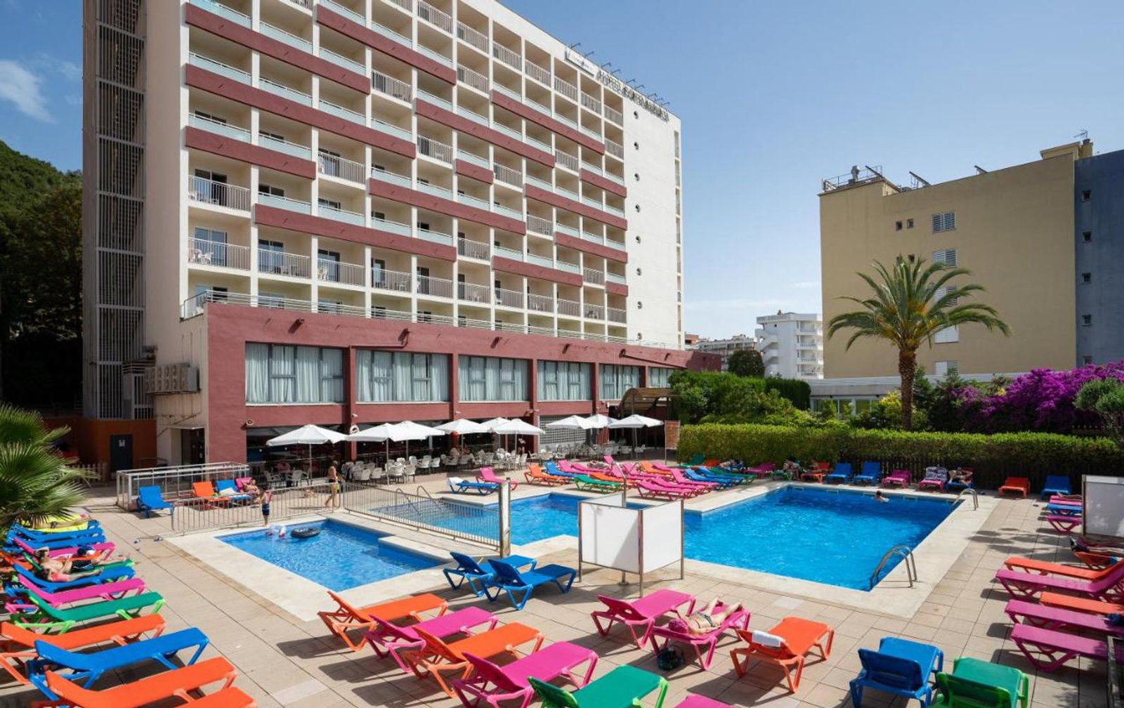Outdoor hotel pool area with colourful sun loungers and parasols in front of a multi-storey building and a palm tree