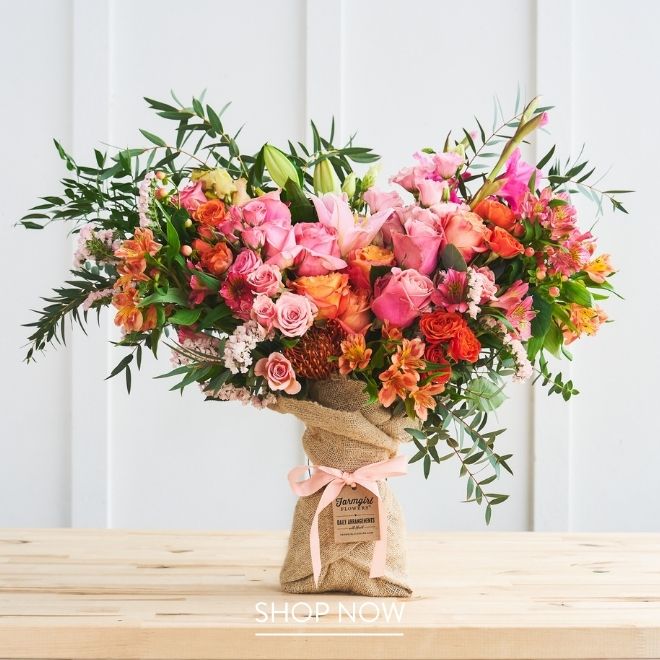 A burlap-wrapped bouquet of pink roses, orange alstroemeria, and pink gladiolus stands centered on the table against a white paneled wall.