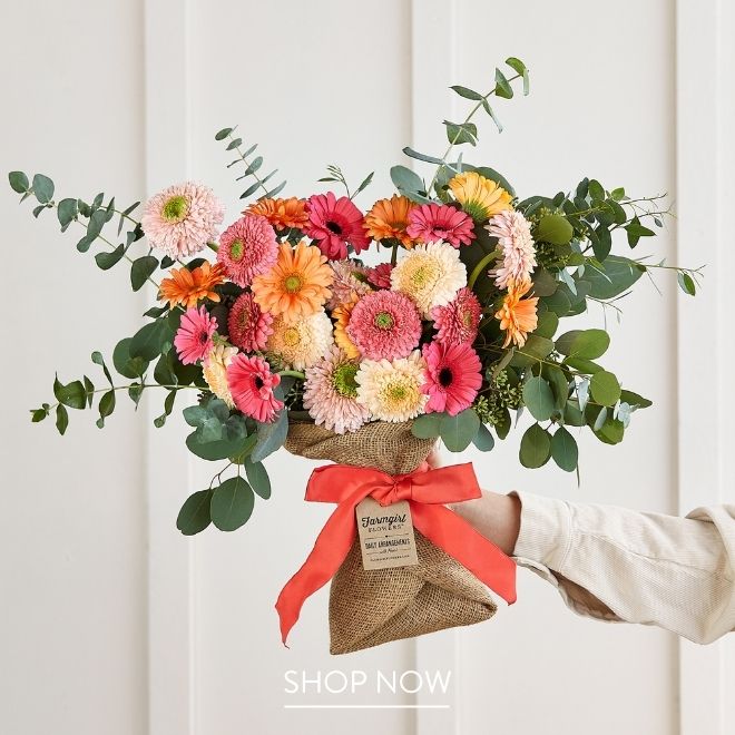 A burlap-wrapped bouquet of pink, coral, and cream gerbera daisies with eucalyptus, tied with a coral ribbon.