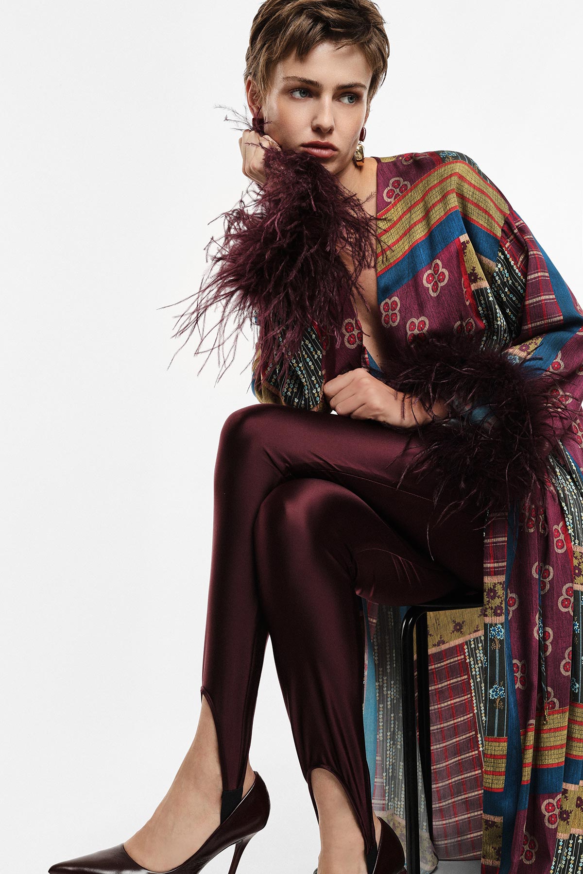 Seated model in patterned kimono coat with feathered cuffs, burgundy satin trousers and heels, posing pensively