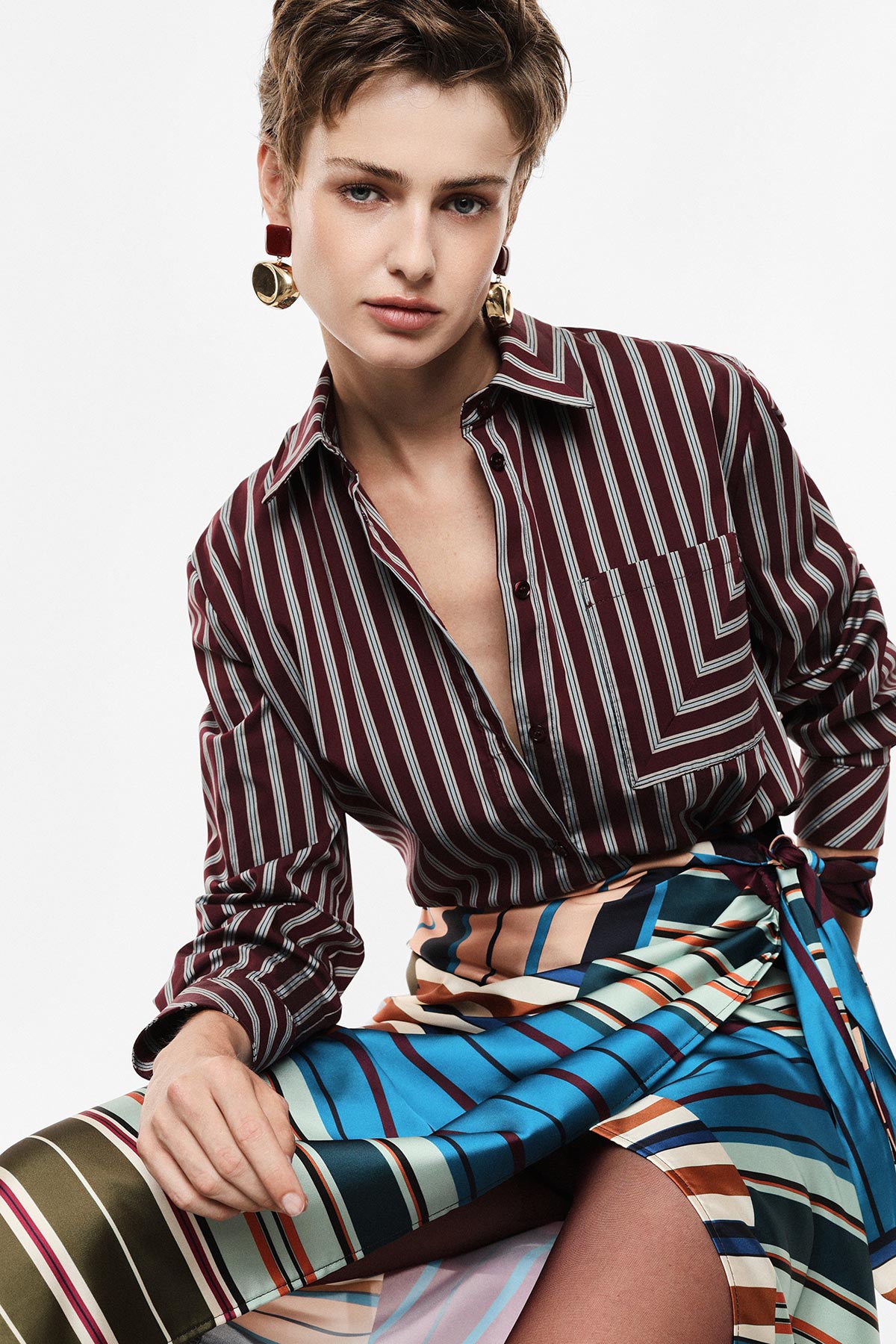 Model wearing burgundy striped shirt and multicolour striped skirt, short hair and gold earrings.