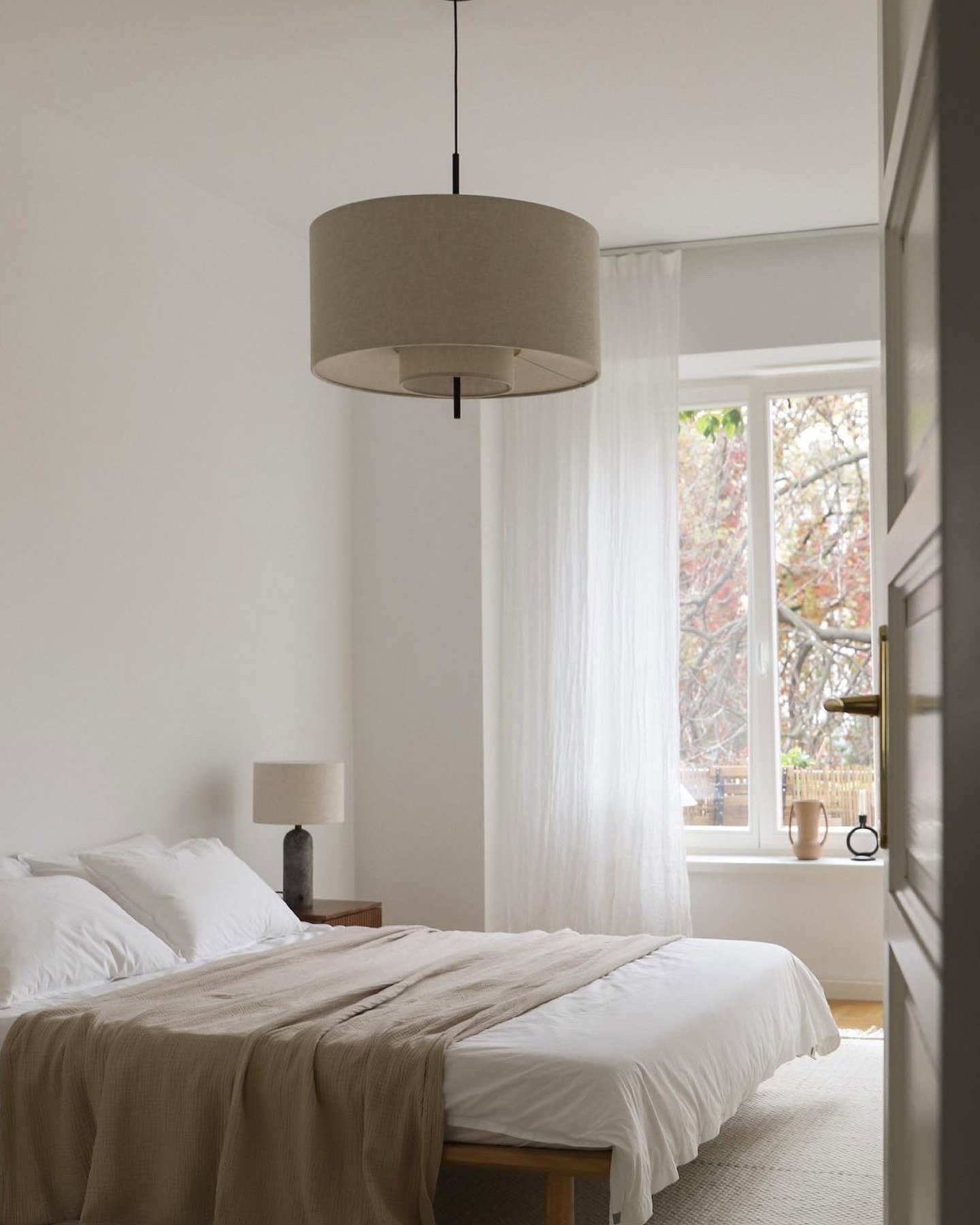 Minimalist neutral-bedroom with white linens, beige throw, pendant light and sheer-curtained window overlooking autumn trees