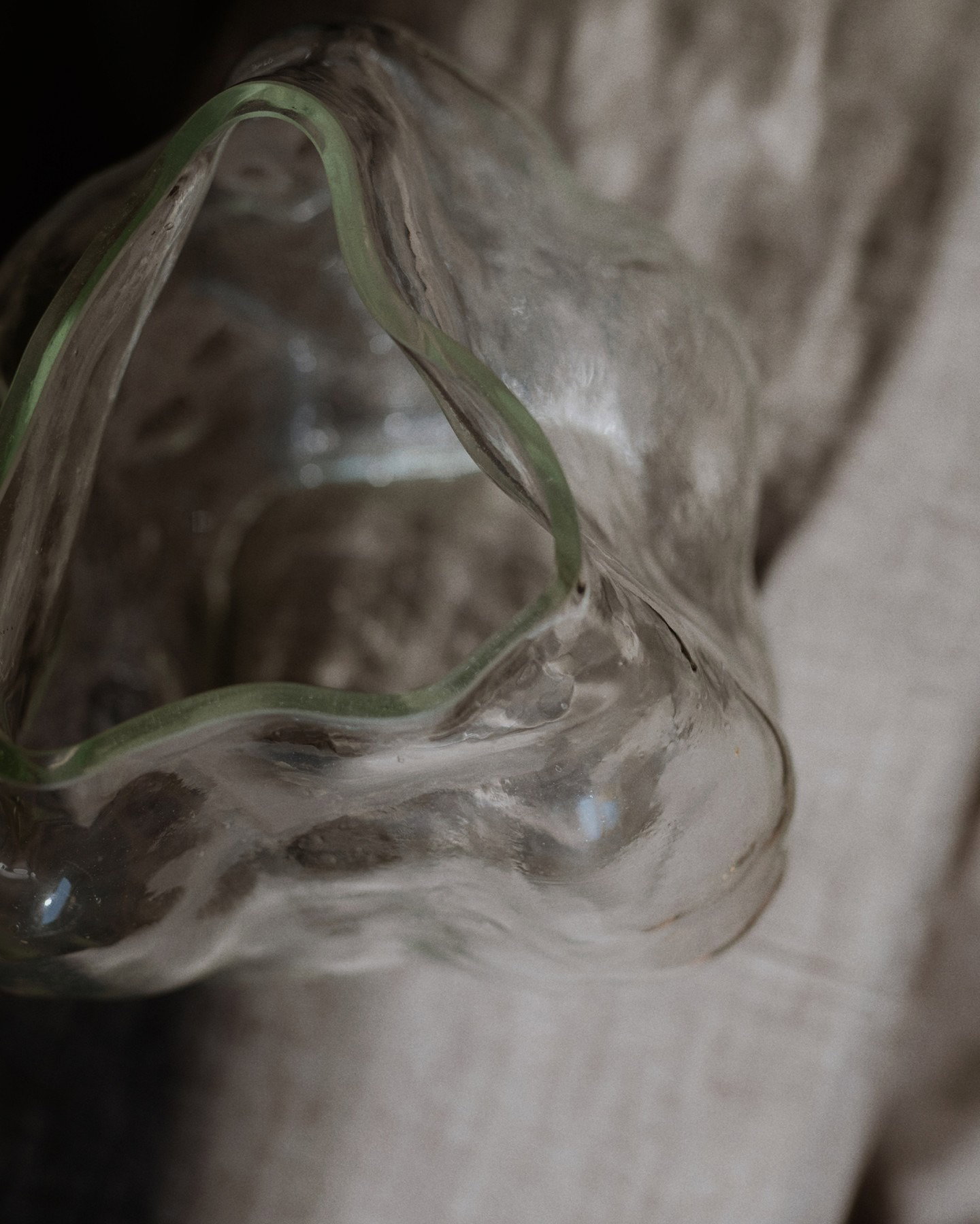 Close-up of a wavy clear glass bowl with a pale green rim resting on textured fabric